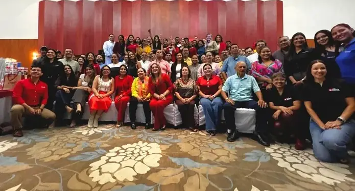 Fotografía grupal de un gran equipo de profesionales sonriendo en un evento corporativo frente a un panel de madera roja.