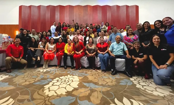 Fotografía grupal de un gran equipo de profesionales sonriendo en un evento corporativo frente a un panel de madera roja.