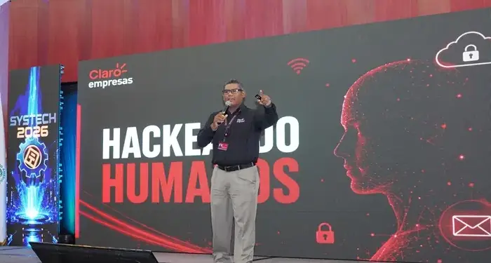 Conferencista en el escenario hablando sobre hackeo humano frente a una gran pantalla con gráficos digitales.