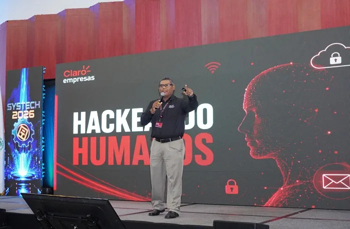 Conferencista en el escenario hablando sobre hackeo humano frente a una gran pantalla con gráficos digitales.