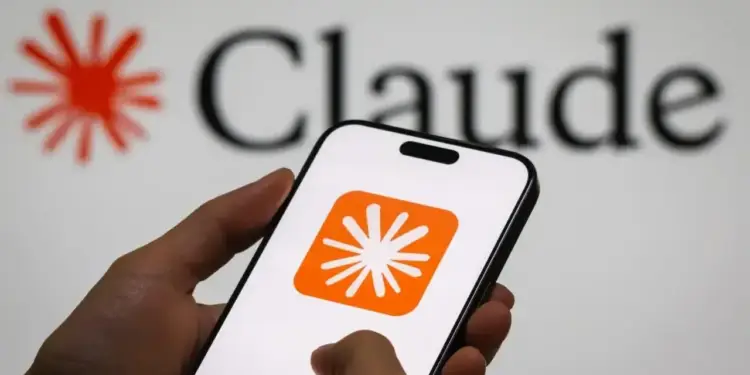 Manos sosteniendo un smartphone blanco que muestra una aplicación con logotipo naranja, con la palabra Claude al fondo.