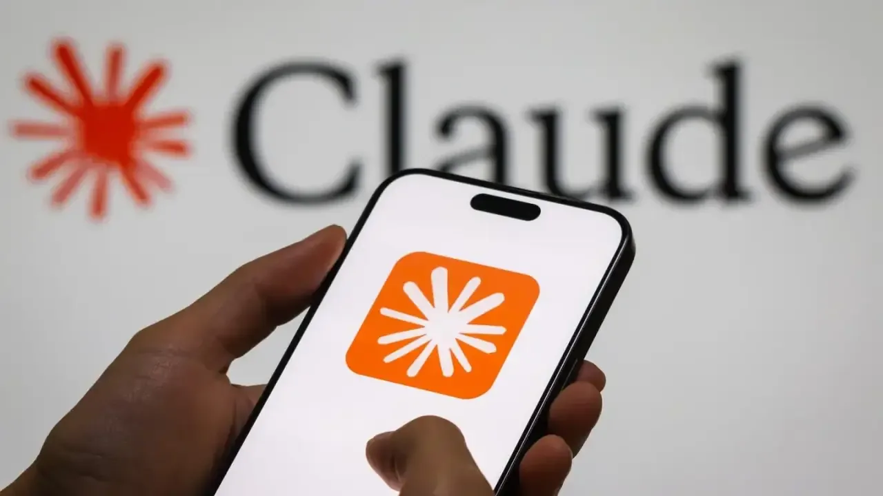 Manos sosteniendo un smartphone blanco que muestra una aplicación con logotipo naranja, con la palabra Claude al fondo.