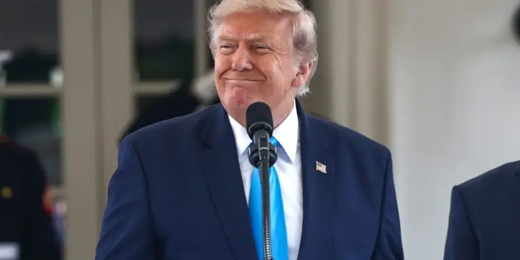 Hombre de cabello rubio con traje azul y corbata celeste sonríe frente a un micrófono. Lleva un pin de la bandera de EE. UU. en la solapa.