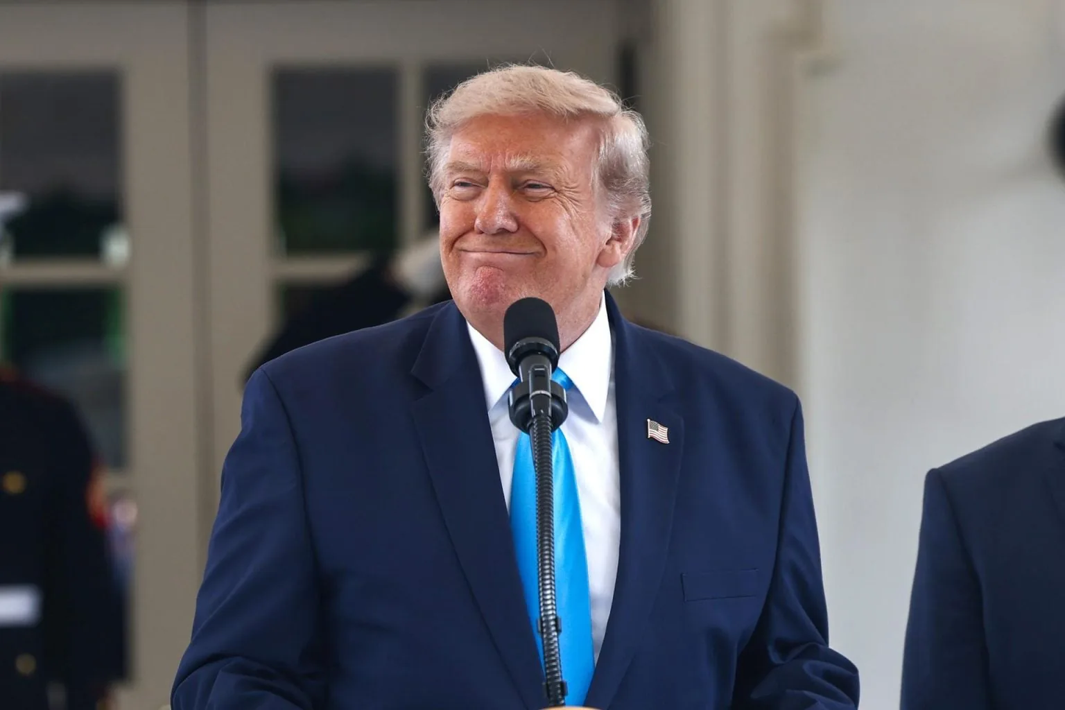 Hombre de cabello rubio con traje azul y corbata celeste sonríe frente a un micrófono. Lleva un pin de la bandera de EE. UU. en la solapa.