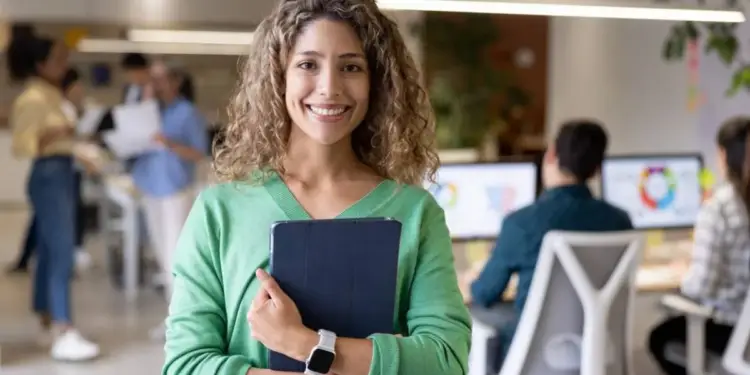Mujer joven y sonriente con cabello rizado vistiendo jersey verde sosteniendo una tablet en una oficina moderna.