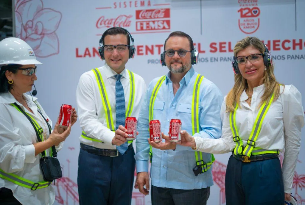 Cuatro ejecutivos con chalecos de seguridad y gafas sostienen latas rojas de refresco en un evento de Coca-Cola FEMSA.