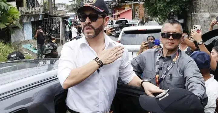 Hombre con camisa blanca, gafas de sol y gorra negra junto a vehículos y personal de seguridad en una calle urbana.