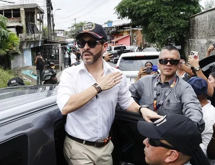 Hombre con camisa blanca, gafas de sol y gorra negra junto a vehículos y personal de seguridad en una calle urbana.