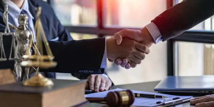 Dos profesionales dándose la mano sobre un escritorio con elementos de justicia como una balanza, libros y un mazo.