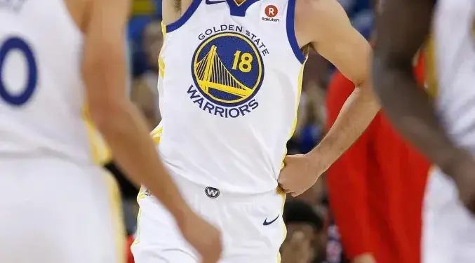 Jugador de baloncesto con el número 18 de Golden State Warriors señala hacia arriba en la cancha durante un partido.