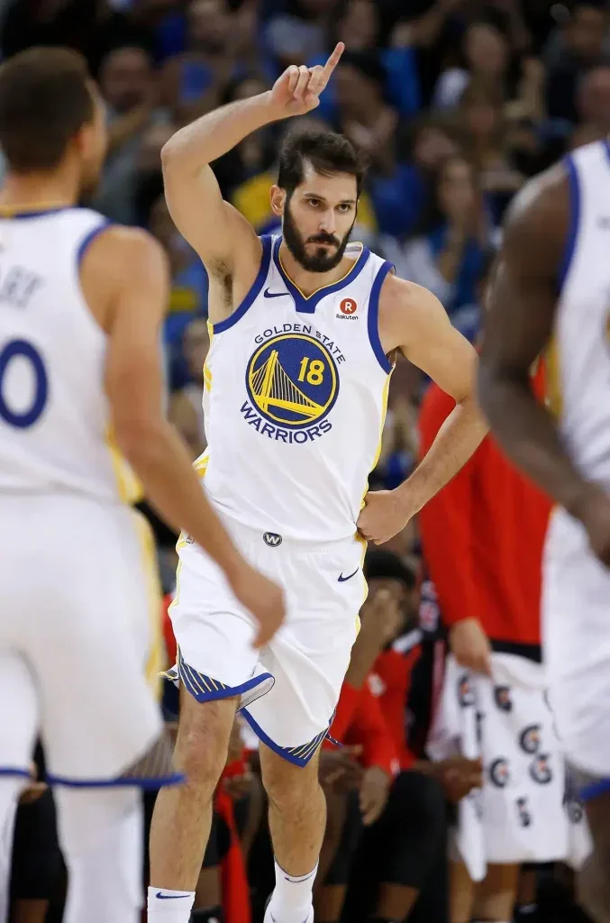 Jugador de baloncesto con el número 18 de Golden State Warriors señala hacia arriba en la cancha durante un partido.