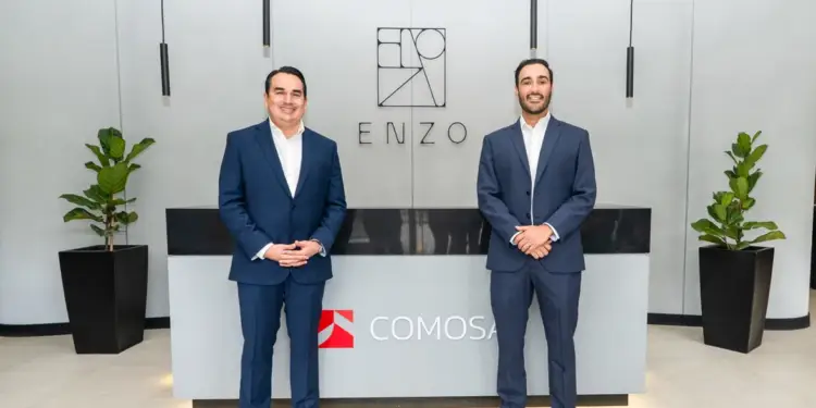 Dos hombres sonrientes con trajes oscuros en una oficina moderna. Están de pie frente a un mostrador de recepción con logos y plantas.