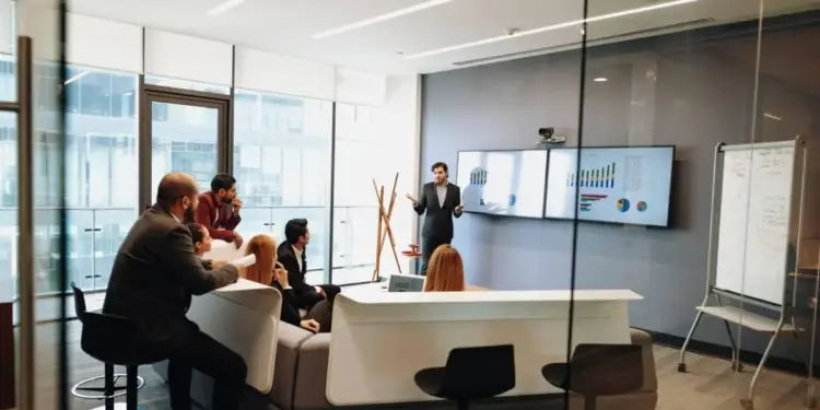 Un grupo de profesionales en una sala de reuniones moderna, escuchando atentamente a un presentador que expone gráficos y datos en pantallas digitales.