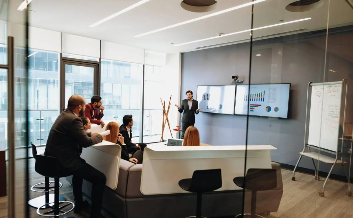 Un grupo de profesionales en una sala de reuniones moderna, escuchando atentamente a un presentador que expone gráficos y datos en pantallas digitales.