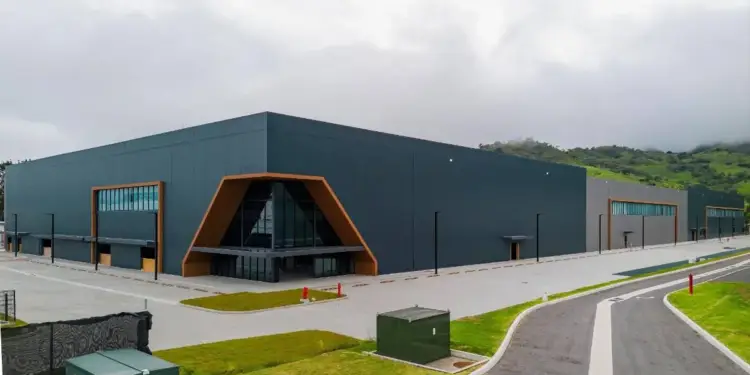 Vista exterior de un edificio industrial moderno con fachada gris oscuro, detalles en madera y entrada principal triangular acristalada, rodeado de áreas verdes y colinas.