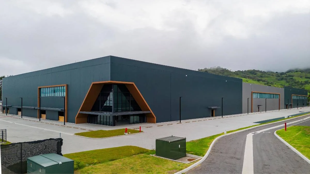 Vista exterior de un edificio industrial moderno con fachada gris oscuro, detalles en madera y entrada principal triangular acristalada, rodeado de áreas verdes y colinas.