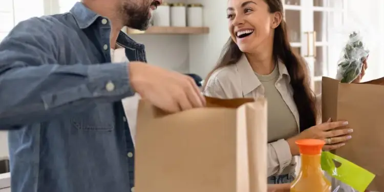 Una pareja sonriente desempaca bolsas de papel con comestibles en la cocina de su hogar.