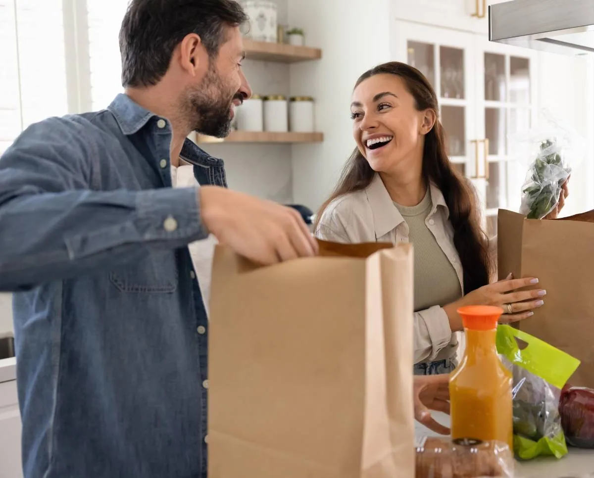 Una pareja sonriente desempaca bolsas de papel con comestibles en la cocina de su hogar.