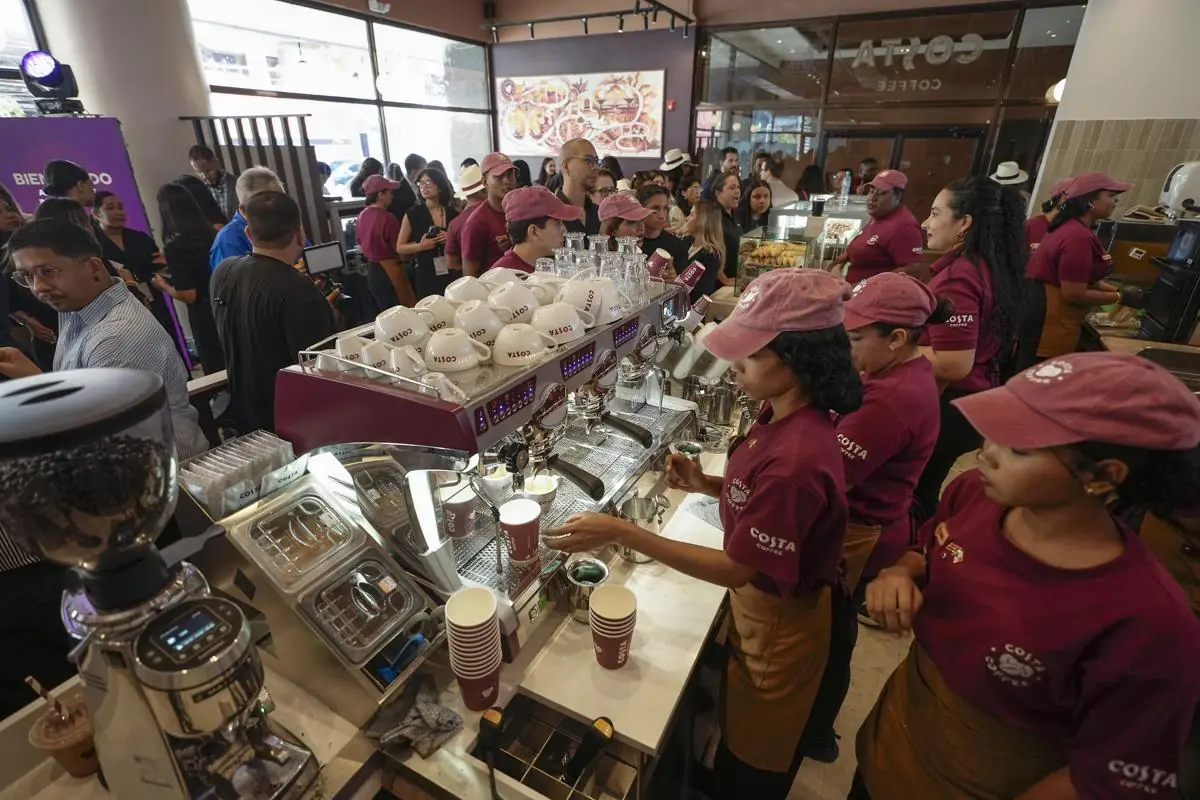 Personal con gorras y uniformes preparando café en una máquina espresso dentro de un local lleno de clientes.