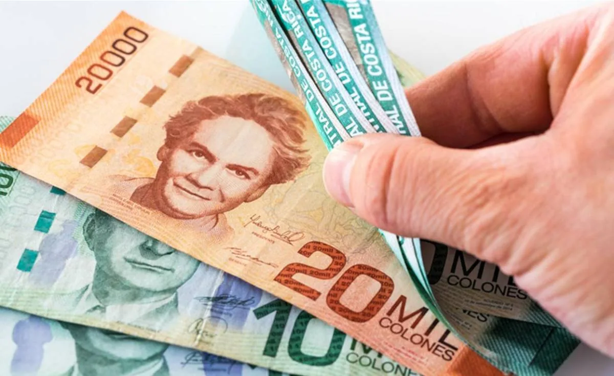 Mano contando billetes de diferentes denominaciones en colones sobre una superficie blanca.