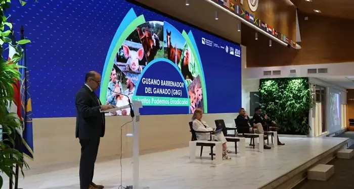 Hombre en podio presenta sobre erradicación del Gusano Barrenador del Ganado en conferencia. Pantalla muestra animales de granja y texto relevante. Panelistas sentados.