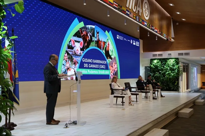 Hombre en podio presenta sobre erradicación del Gusano Barrenador del Ganado en conferencia. Pantalla muestra animales de granja y texto relevante. Panelistas sentados.