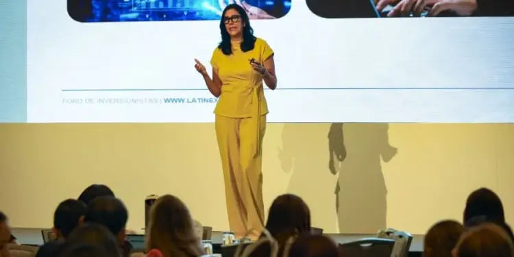 Mujer experta en finanzas y tecnología presenta en un foro de inversión. La pantalla muestra gráficos financieros, un brazo robótico y datos digitales. Audiencia atenta.