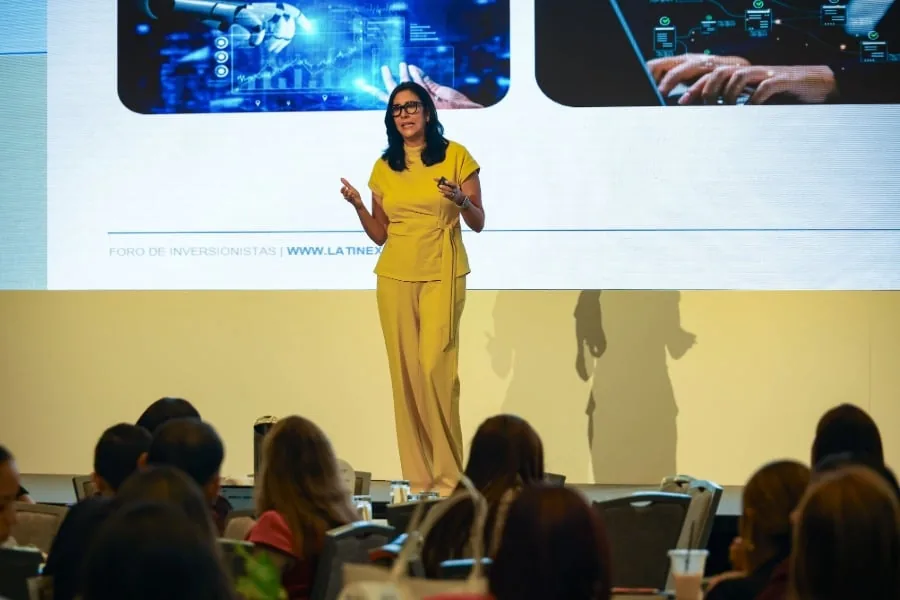 Mujer experta en finanzas y tecnología presenta en un foro de inversión. La pantalla muestra gráficos financieros, un brazo robótico y datos digitales. Audiencia atenta.