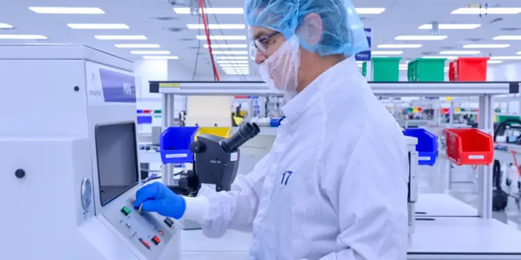 Hombre con bata de laboratorio, gorro y mascarilla operando una máquina de control de calidad en una sala limpia. Sostiene un componente con código de barras.