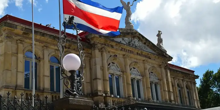 Bandera de Costa Rica ondeando en un asta frente a la fachada arquitectónica clásica del Teatro Nacional bajo un cielo nublado.