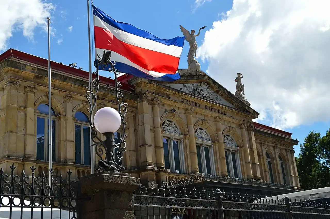 Bandera de Costa Rica ondeando en un asta frente a la fachada arquitectónica clásica del Teatro Nacional bajo un cielo nublado.