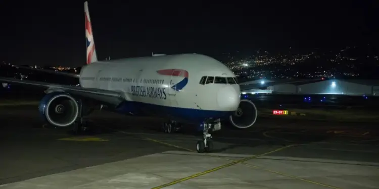 Gran avión de pasajeros British Airways, blanco y azul, en la pista de un aeropuerto por la noche, con luces urbanas al fondo.