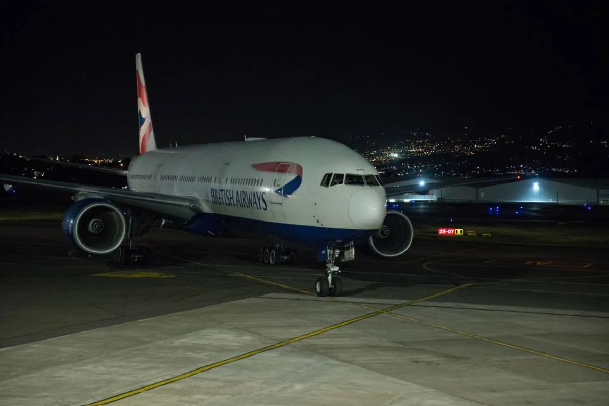 Gran avión de pasajeros British Airways, blanco y azul, en la pista de un aeropuerto por la noche, con luces urbanas al fondo.
