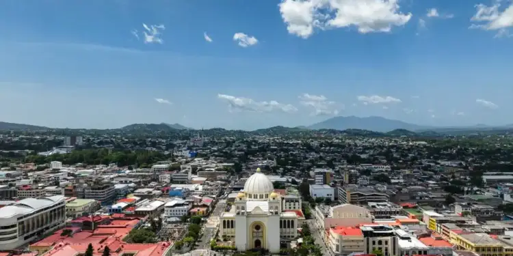 Vista aérea de una ciudad con un edificio central de cúpula blanca, rodeado de edificios, calles y montañas al fondo bajo un cielo azul claro.