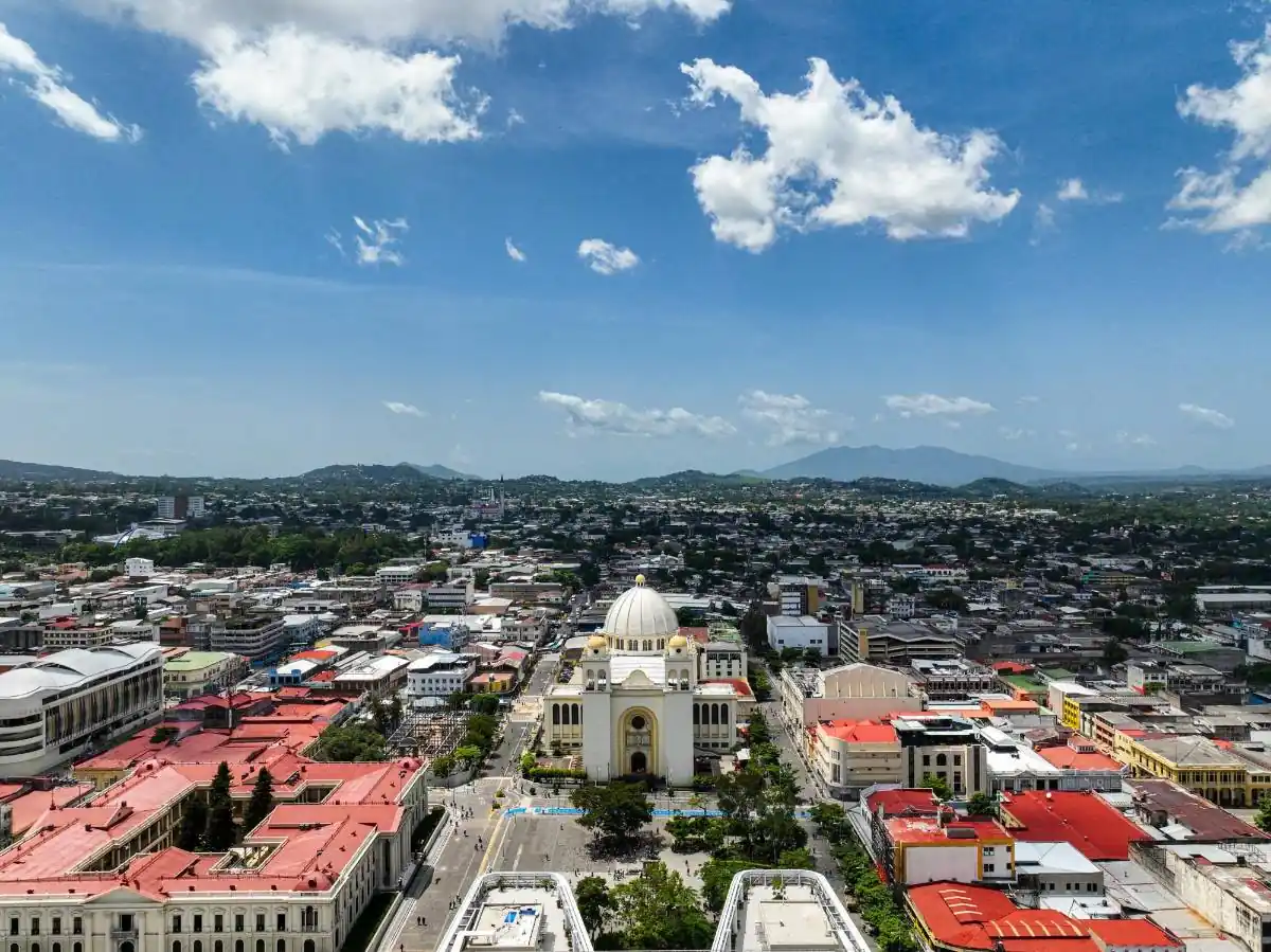 Vista aérea de una ciudad con un edificio central de cúpula blanca, rodeado de edificios, calles y montañas al fondo bajo un cielo azul claro.