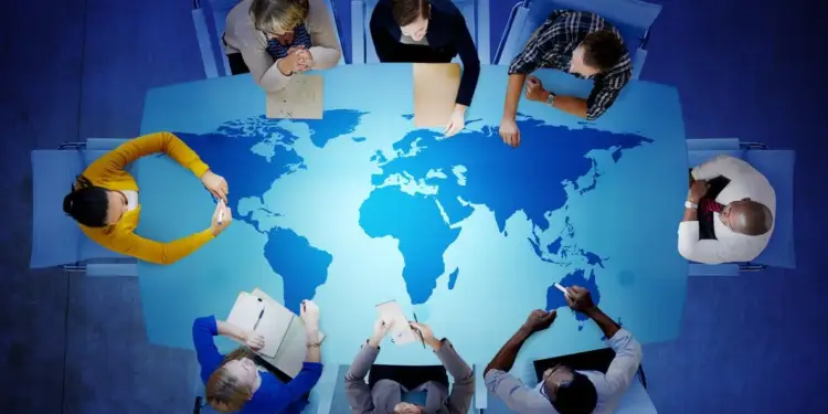 Vista aérea de ocho profesionales de negocios sentados alrededor de una mesa con un mapa mundial. Simboliza estrategia global y colaboración internacional.