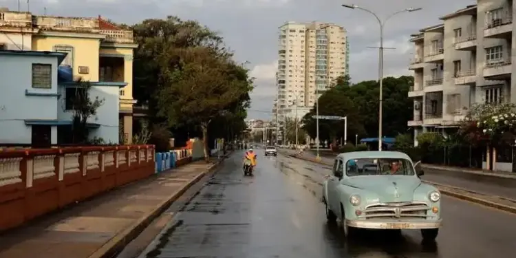 Vista de una calle urbana mojada por la lluvia, con un auto clásico celeste en primer plano, otros vehículos y edificios de diferentes épocas al fondo.
