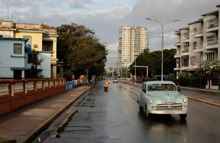 Vista de una calle urbana mojada por la lluvia, con un auto clásico celeste en primer plano, otros vehículos y edificios de diferentes épocas al fondo.