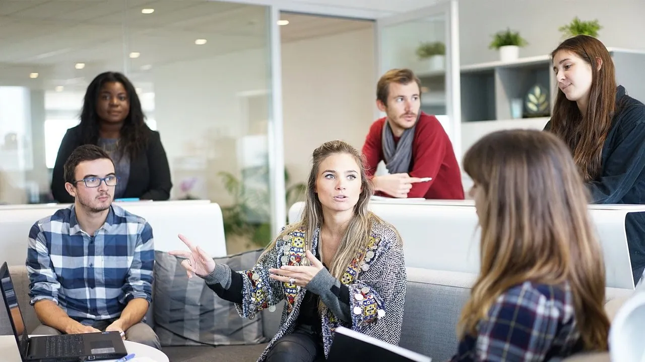 Grupo diverso de profesionales en una reunión de negocios, discutiendo ideas en un entorno colaborativo de oficina moderna. Una mujer gesticula mientras habla.
