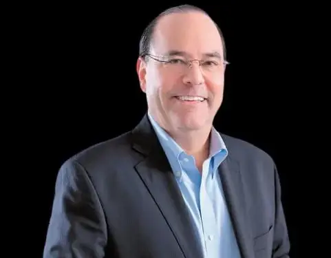Retrato de un hombre caucásico de mediana edad, sonriente, con gafas, vistiendo un traje oscuro y camisa azul claro, sobre fondo negro. Representa profesionalismo.