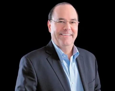Retrato de un hombre caucásico de mediana edad, sonriente, con gafas, vistiendo un traje oscuro y camisa azul claro, sobre fondo negro. Representa profesionalismo.