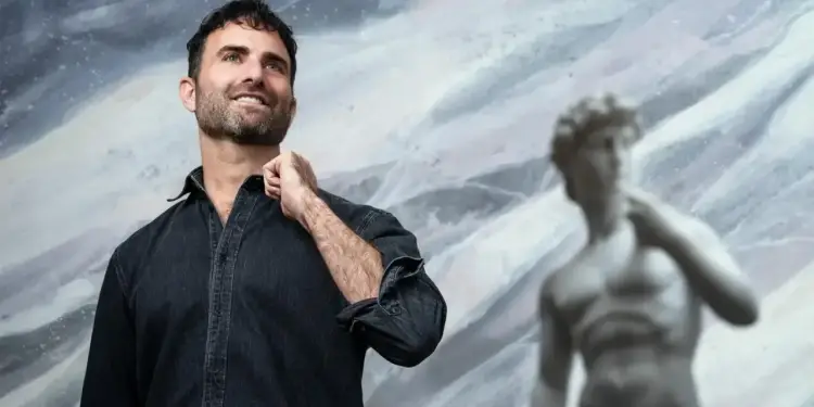 Hombre barbudo sonriente con camisa de mezclilla oscura, mirando hacia arriba, con una estatua borrosa de David de fondo abstracto.