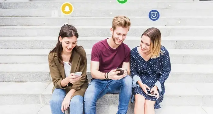 Tres jóvenes sentados en unas escaleras de piedra interactuando con sus teléfonos móviles, rodeados de iconos de redes sociales flotantes.