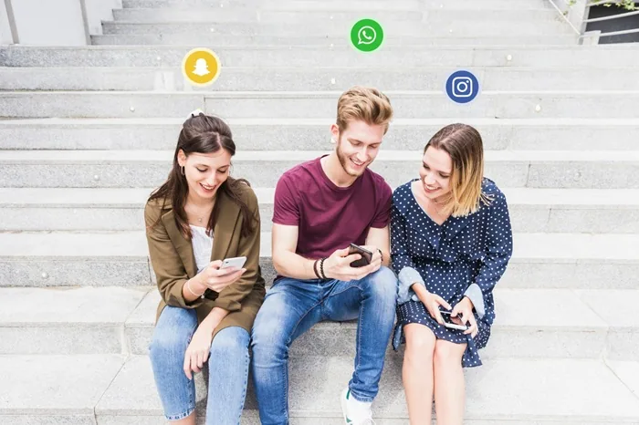 Tres jóvenes sentados en unas escaleras de piedra interactuando con sus teléfonos móviles, rodeados de iconos de redes sociales flotantes.
