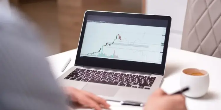 Persona analizando gráficos financieros y de bolsa en la pantalla de una laptop sobre un escritorio blanco.