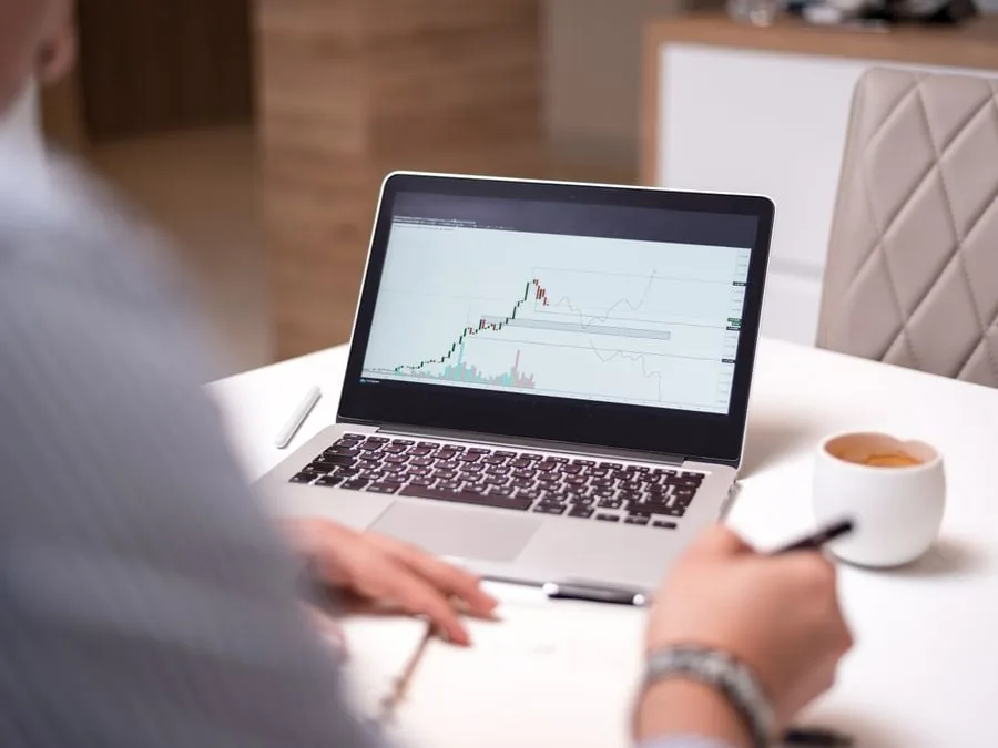Persona analizando gráficos financieros y de bolsa en la pantalla de una laptop sobre un escritorio blanco.