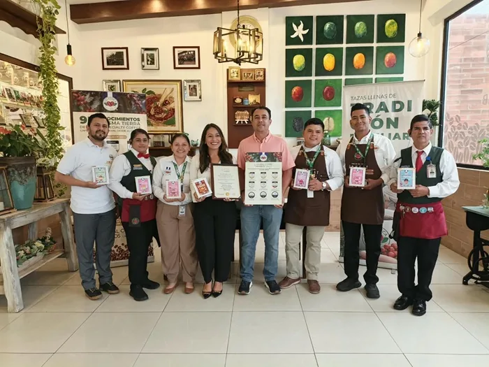 Ocho profesionales, hombres y mujeres, posan con certificados y productos de café. Visten uniformes y ropa de negocios, celebrando logros empresariales y calidad.