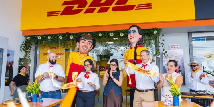 Grupo de personas cortando una cinta amarilla frente a una sucursal de DHL con mojigangas gigantes.