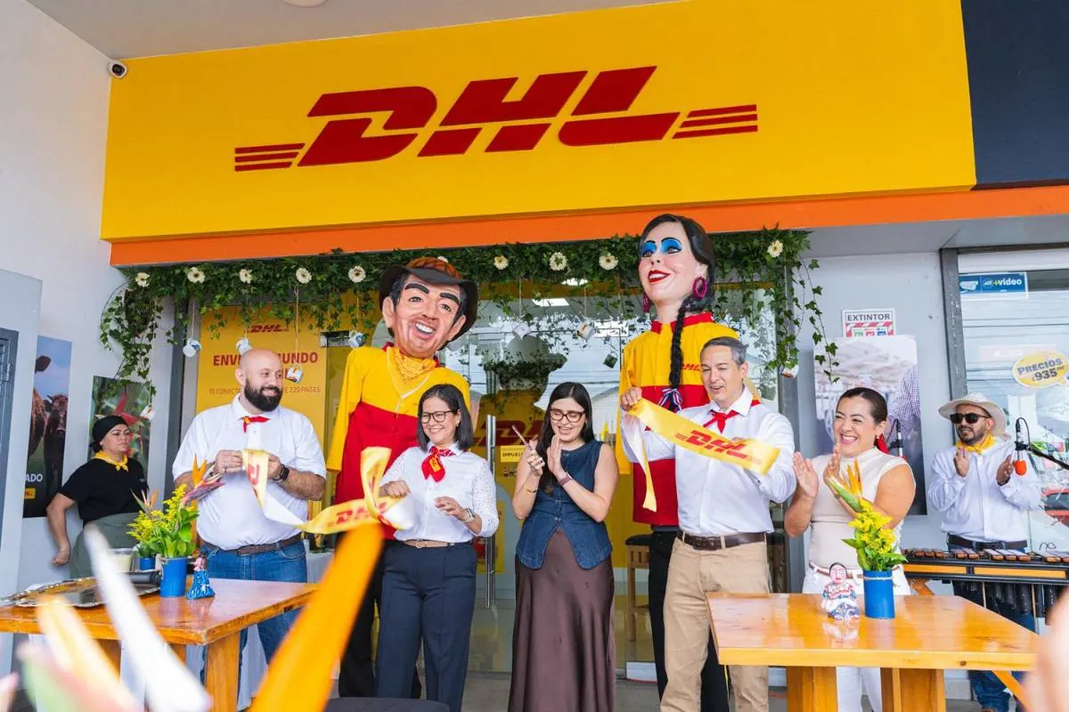 Grupo de personas cortando una cinta amarilla frente a una sucursal de DHL con mojigangas gigantes.