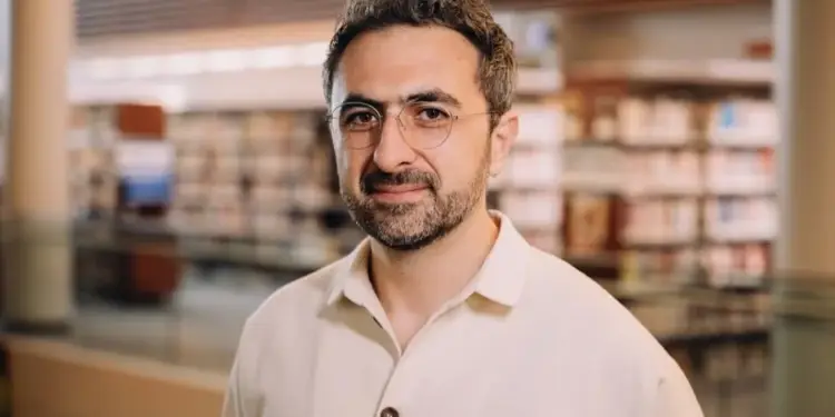 Hombre con gafas y camisa clara sonriendo en un espacio interior con estanterías de fondo.